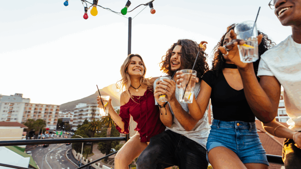 Group of young friends on a rooftop at sunset, laughing and holding drinks.