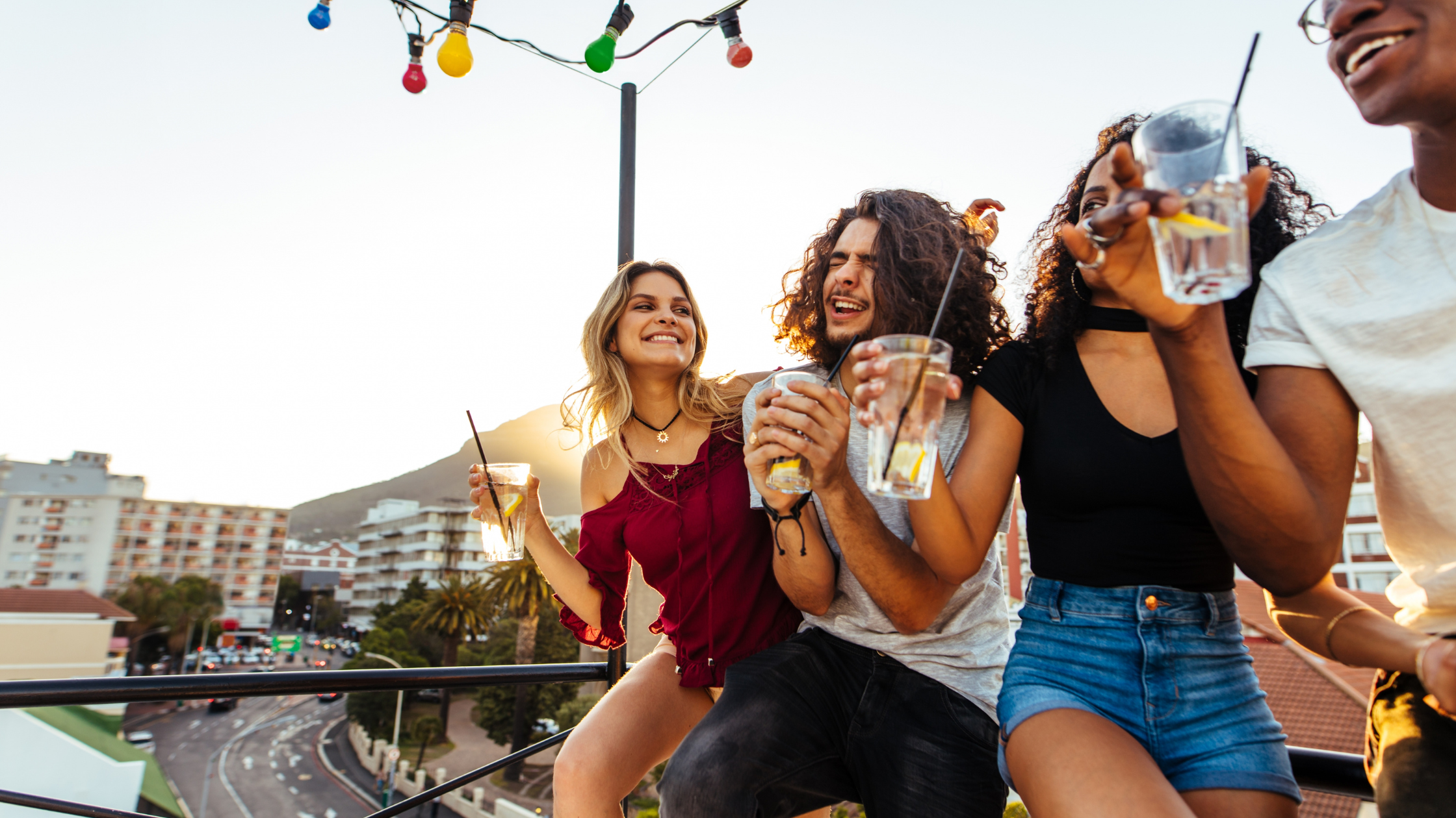 Group of young friends on a rooftop at sunset, laughing and holding drinks.