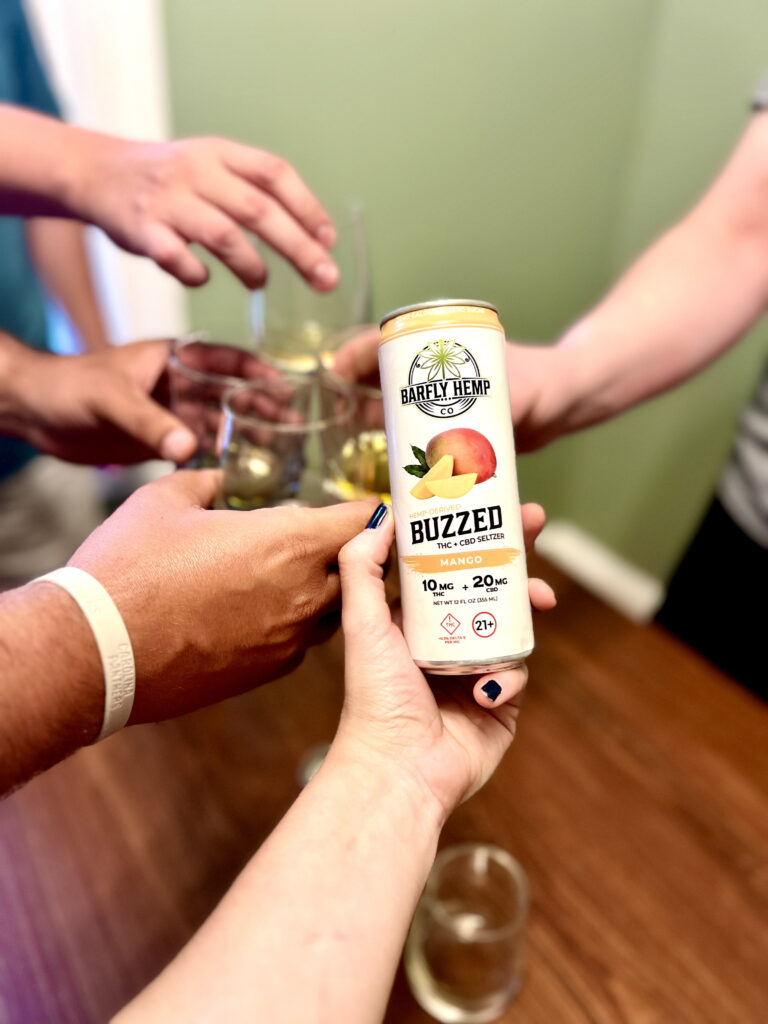 People clinking glasses in a room with a Barfly Mango Buzzed Seltzer held in the foreground