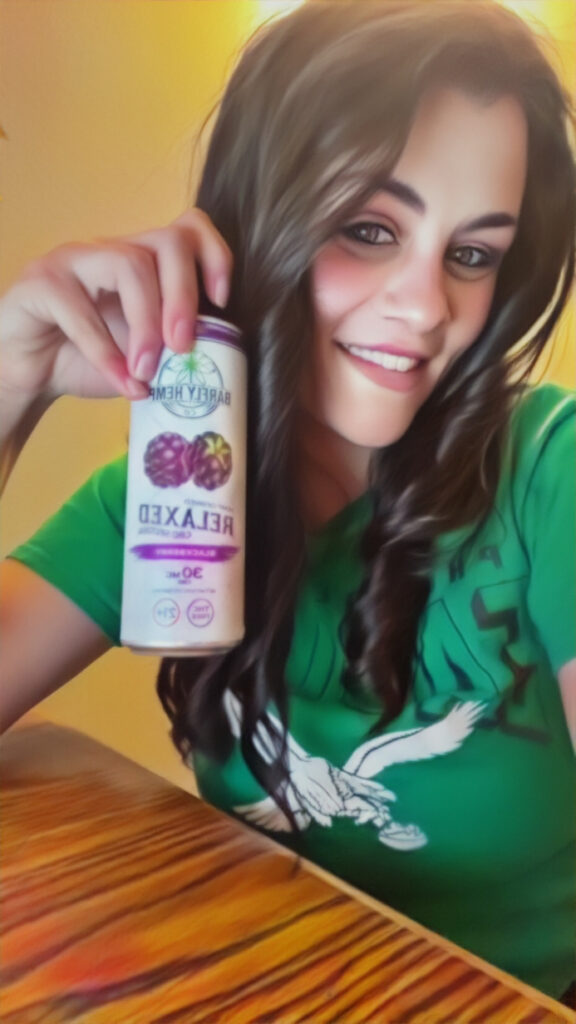 A woman with long dark hair smiles while holding a can of Barfly Hemp Co. Relaxed Blackberry CBD seltzer. She is wearing a green shirt and sitting at a wooden table with a warm yellow background.