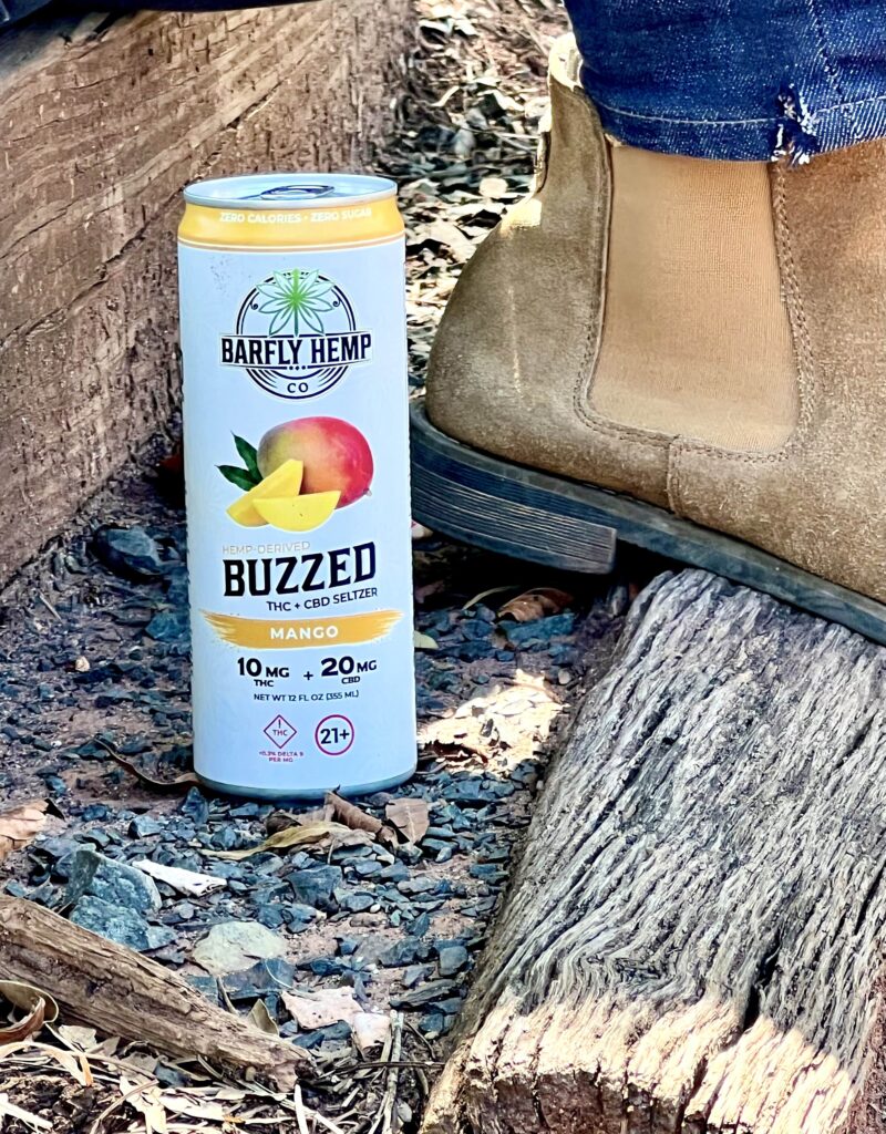 Close-up of a Barfly Hemp Co. Buzzed Mango THC + CBD seltzer can resting on outdoor wooden steps beside tan suede boots.