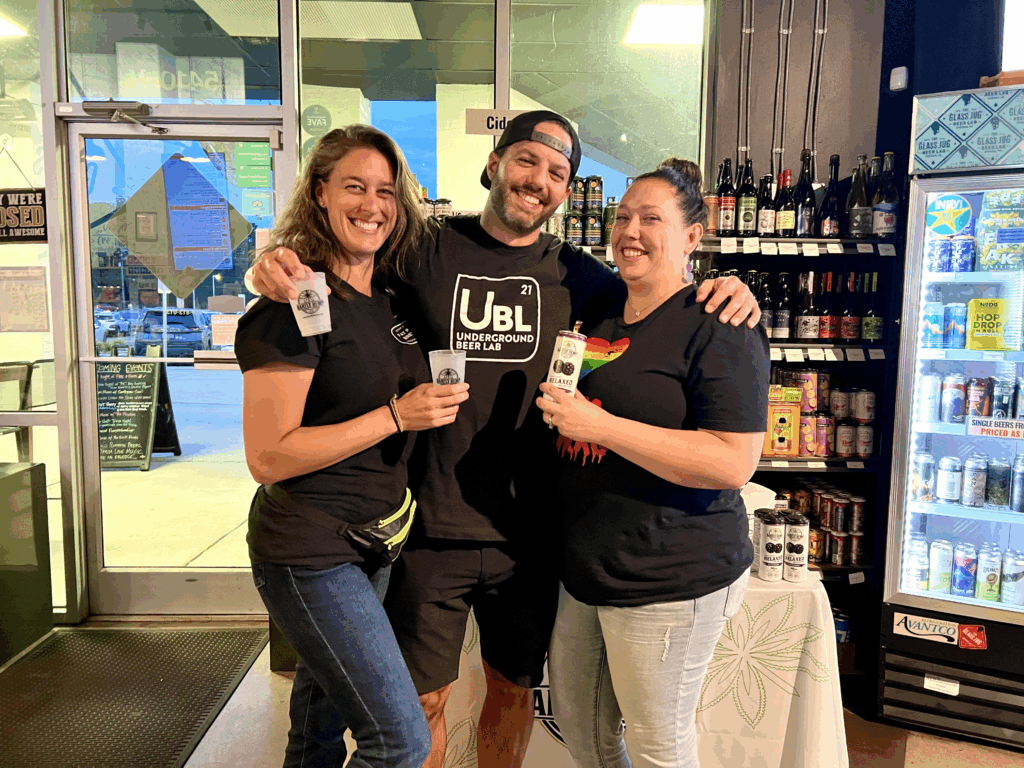 Three smiling people stand together inside a bottle shop during a Barfly Hemp Co. tasting event. Two hold sample cups while one holds a can of Relaxed Blackberry CBD seltzer, with shelves of drinks and a cooler in the background.