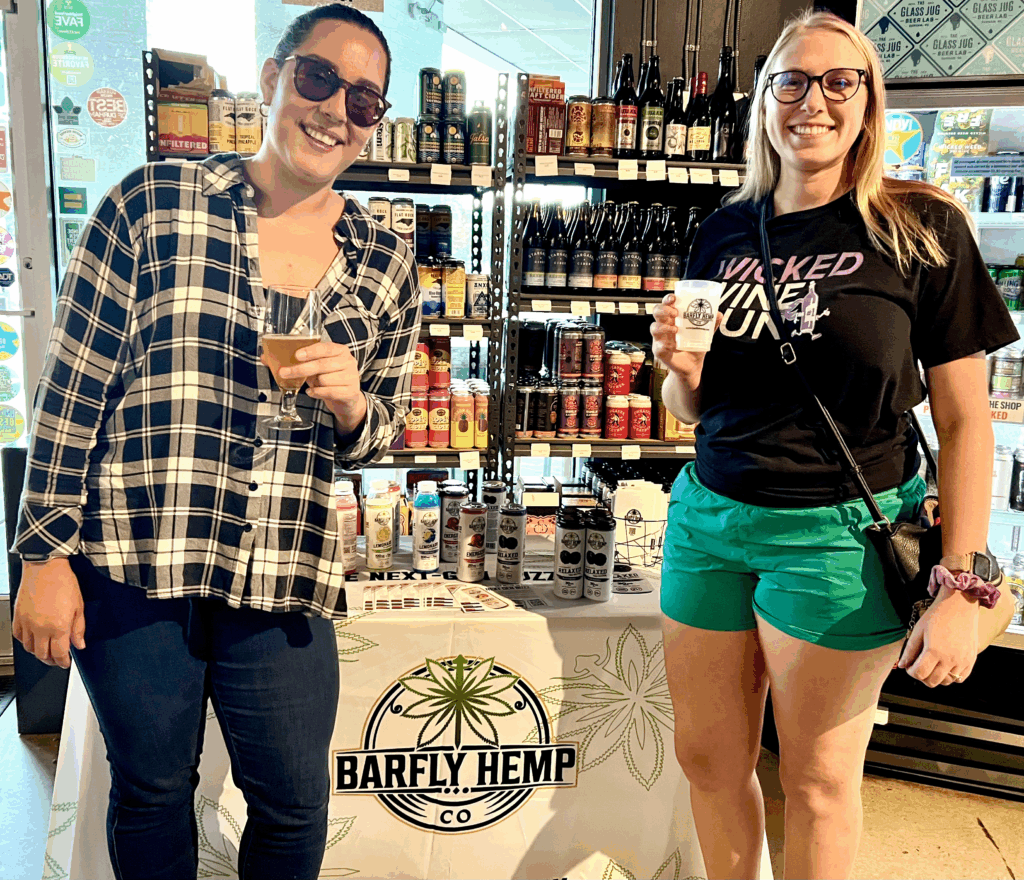 Two women smile while holding Barfly Hemp Co. samples at a store tasting table. Behind them, shelves of craft drinks and a display of Barfly seltzers are visible.
